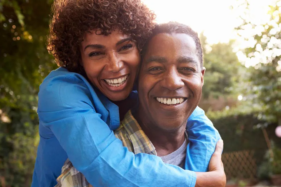 Autoimmune-Doctor A middle-aged African American couple outside, the woman embracing the man from behind pressing her cheek to his, both are smiling due to successful Autoimmune Disorder treatment from Better Life Carolinas of Mount Pleasant in Charleston Metro.