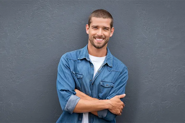 Hormones-for-Men-Doctor A man in a denim shirt stands smiling against a gray-blue wall, pleased with his testosterone hormone therapy from Better Life Carolinas of Mount Pleasant in Charleston Metro.