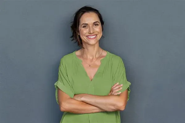 Hormones-for-Women-Doctor A woman in a green shirt stands smiling against a gray-blue wall, pleased with her hormone therapy from Better Life Carolinas of Mount Pleasant in Charleston Metro.
