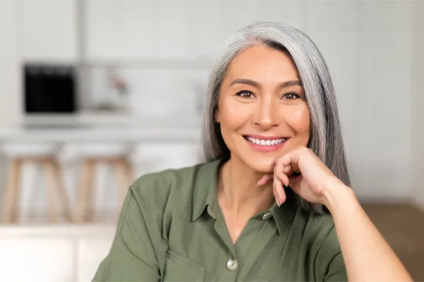 Menopause-Clinic An asian woman with long gray hair and a green shirt sitting and smiling on her couch, happy with her menopause care from [NAME} in Charleston Metro.