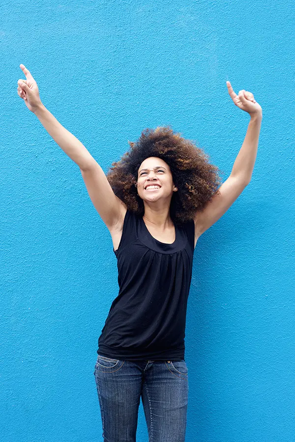 PMS-Treatment A woman in a dark blue tank top standing in front of a bright blue wall, raising her arms in celebration of relief from PMS from Better Life Carolinas of Mount Pleasant in Charleston Metro.