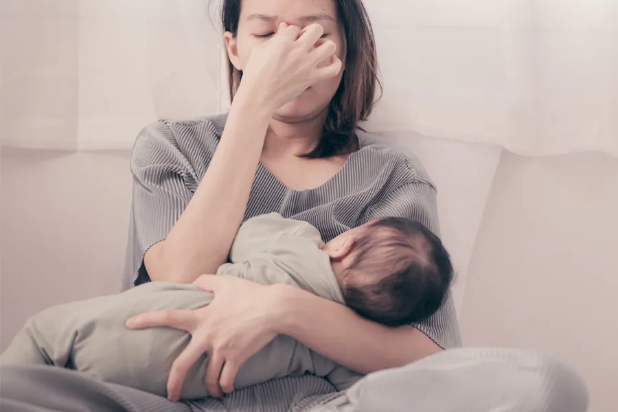 Postpartum-Hormone-Imbalance-Clinic A woman nursing an infant on couch rubs her eyes with exhaustion. Get treatment for postpartum hormone imbalance from Better Life Carolinas of Mount Pleasant in Charleston Metro.